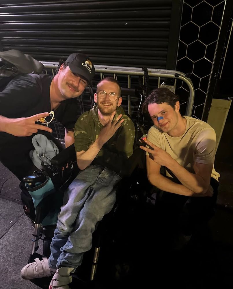 Late at night, Tom with two mates crouched either side of his powerchair, all three throwing up peace signs.